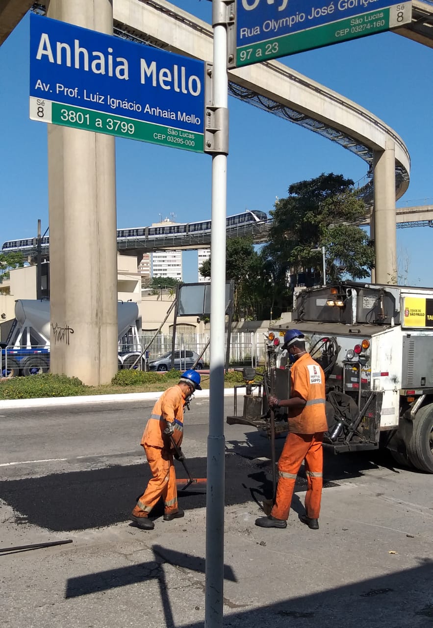 Foto mostra funcionários da Subprefeitura realizando o serviço de tapa buracos na Avenida Anhaia Mello, com a placa do nome da rua em primeiro plano e o Monotrilho ao fundo